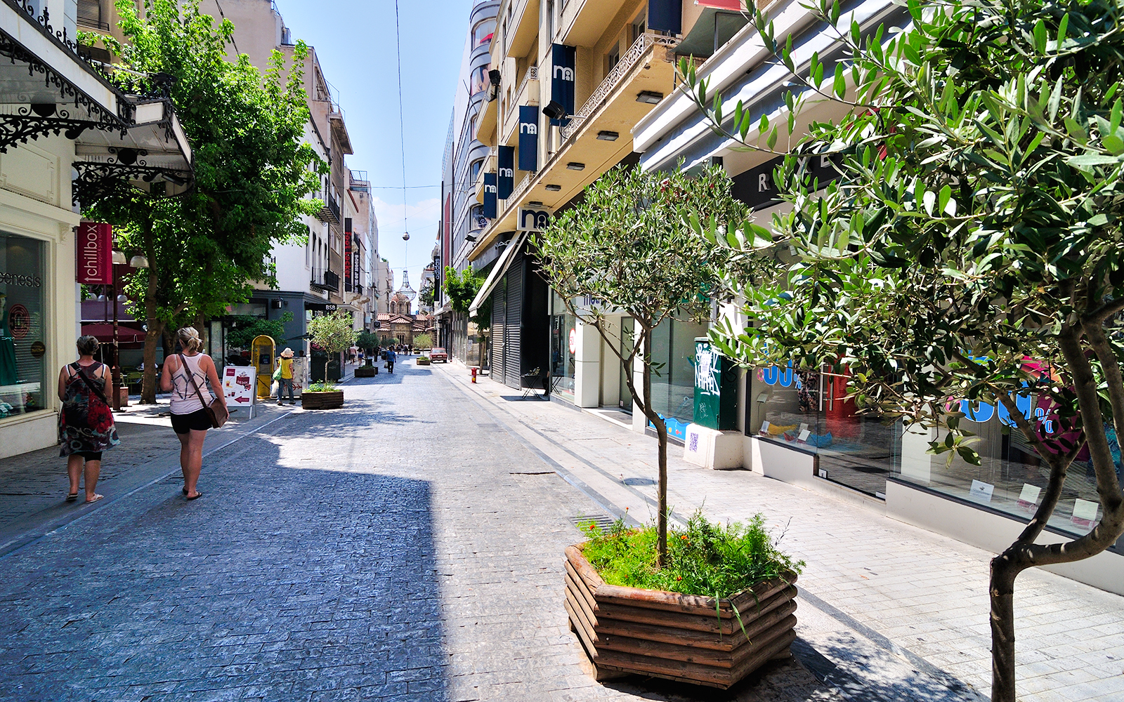 Pedestrians walking along Ermou Street, Athens, lined with shops and trees.