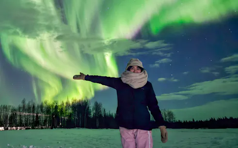 Person enjoying Northern Lights in Kiruna, Sweden, with snow-covered landscape and vibrant aurora.