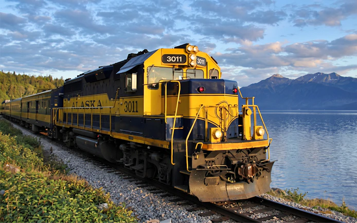 Alaska Railroad train traveling along a scenic lakeside route with mountains in the background.