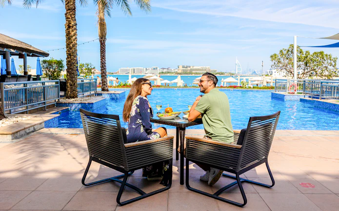 Couple dining by the pool at Riva Beach Club with city skyline view.