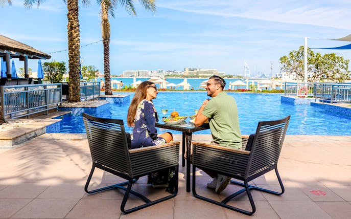 Couple dining by the pool at Riva Beach Club with city skyline view.