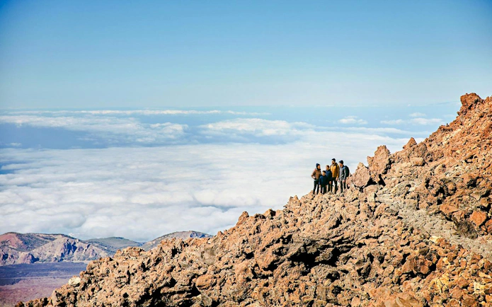 Group of hikers on rocky Mount Teide summit with clouds below, Tenerife.