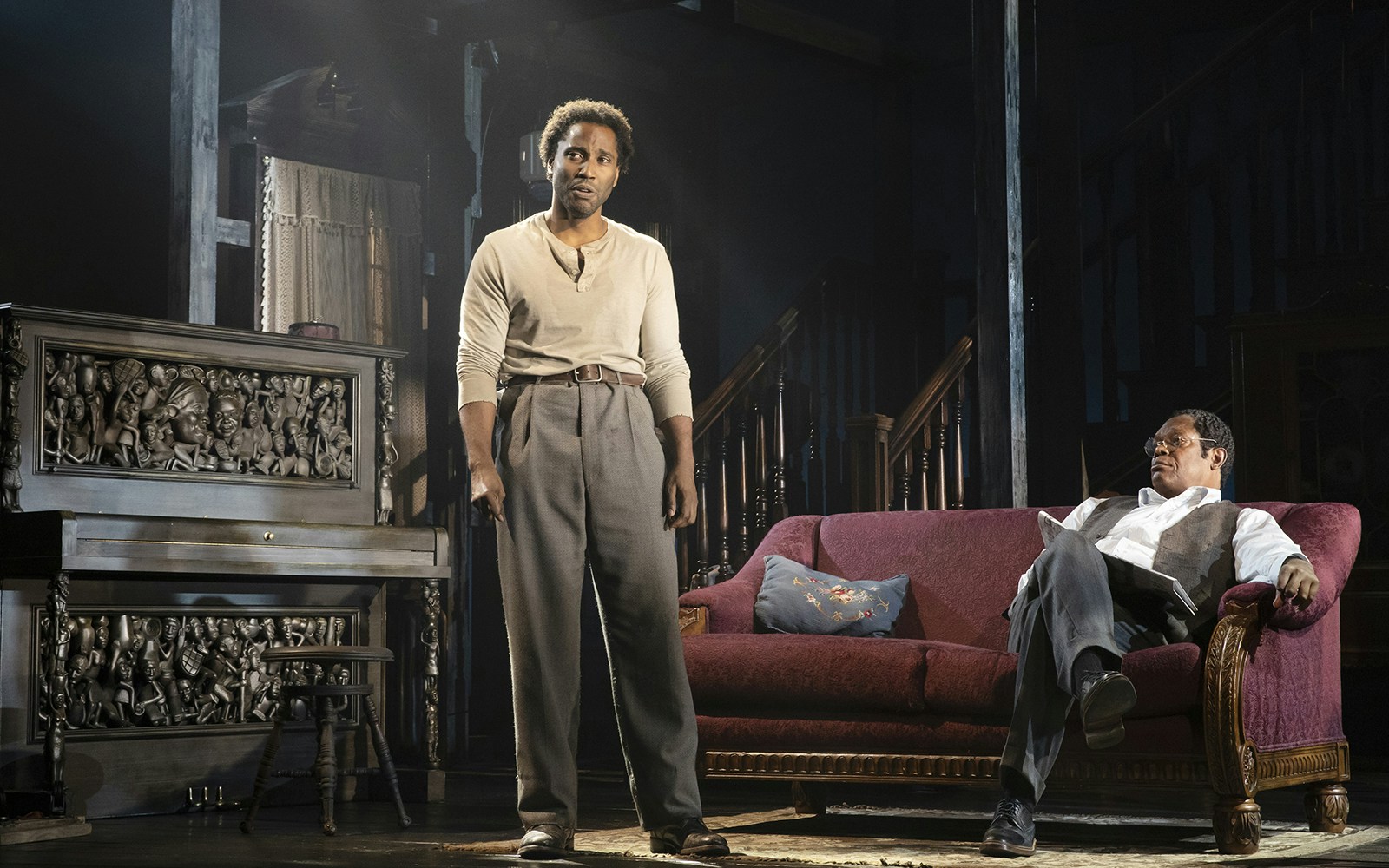 Two actors perform a scene from "The Piano Lesson" with a carved piano and vintage sofa.