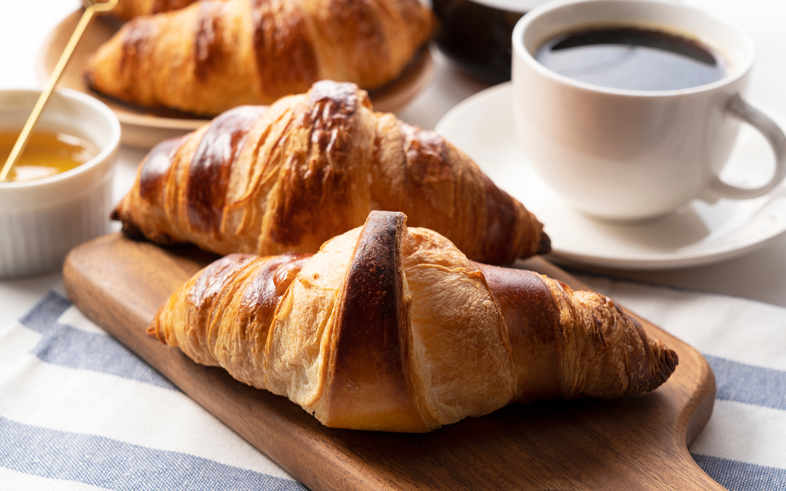 A croissant, and coffee and honey behind it in a cup