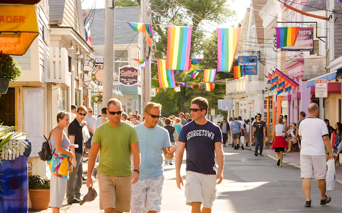 People walking on a colorful street in Provincetown, Cape Cod, with rainbow flags overhead.
