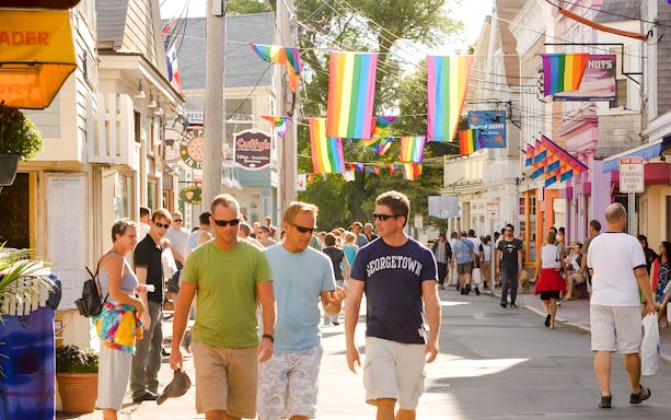 People walking on a colorful street in Provincetown, Cape Cod, with rainbow flags overhead.