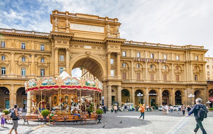 Carousel in Republic Square, Florence, with historic arch and people walking nearby.