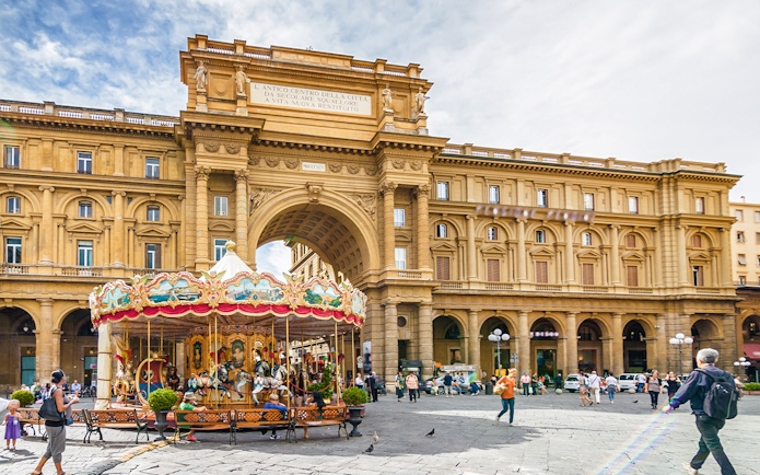 Carousel in Republic Square, Florence, with historic arch and people walking nearby.