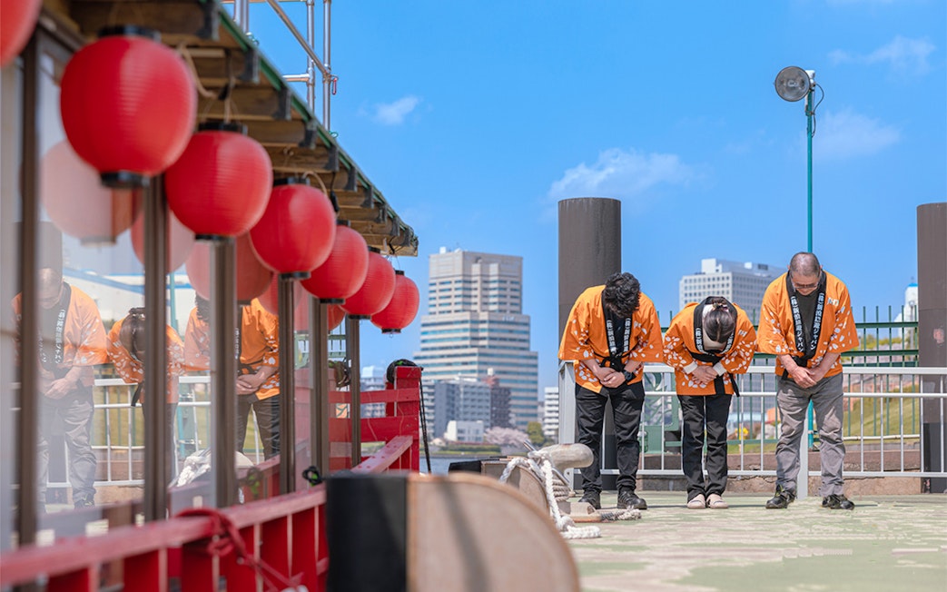 Yakatabune crew in traditional attire bowing on deck with red lanterns in Tokyo.