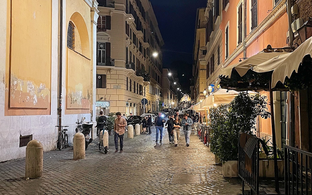 Cobblestone street in Rome at night with people walking and outdoor cafes.