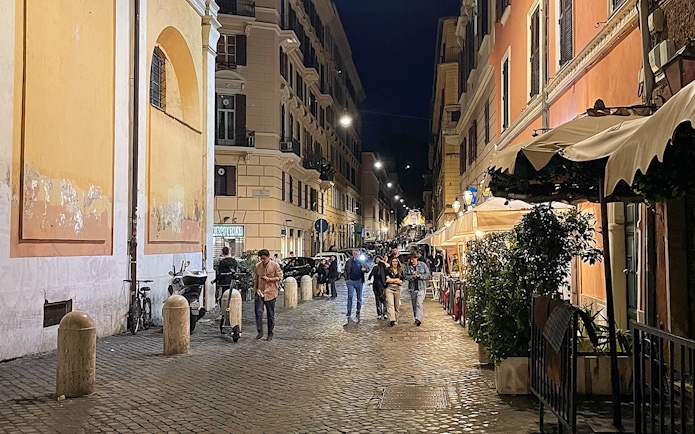 Cobblestone street in Rome at night with people walking and outdoor cafes.