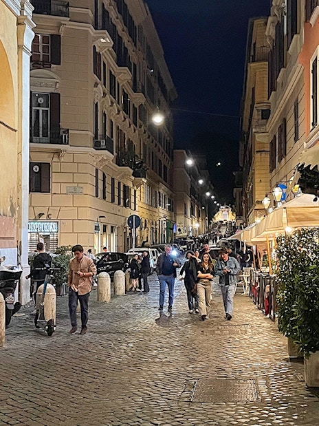Cobblestone street in Rome at night with people walking and outdoor cafes.