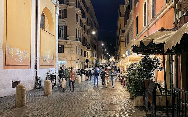 Cobblestone street in Rome at night with people walking and outdoor cafes.