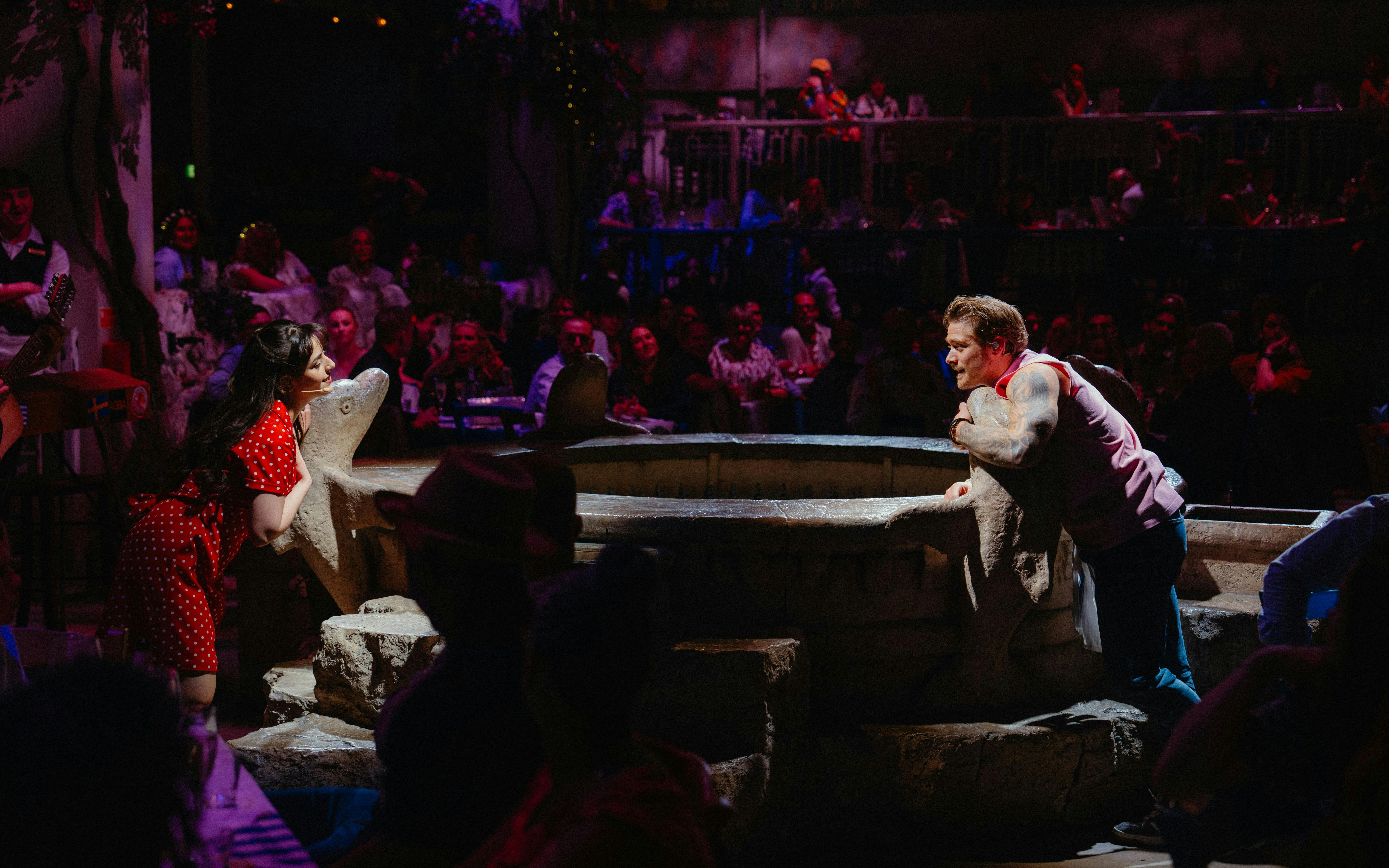 Actors performing a scene from Mamma Mia! The Party with an audience in the background.