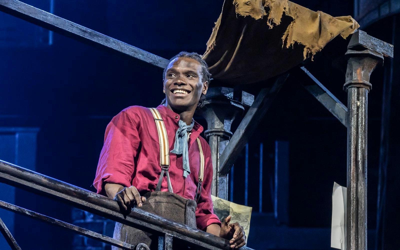 Performer in Newsies musical on stage, wearing period costume, leaning on railing.