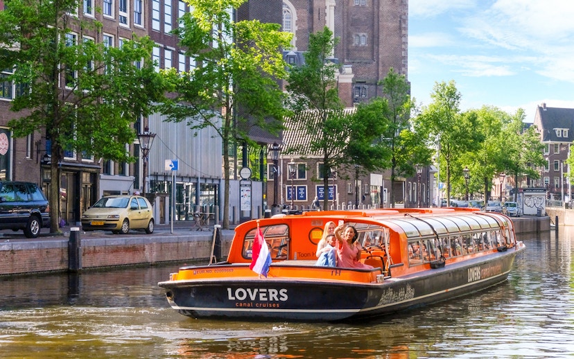 Amsterdam canal cruise boat with tourists waving, passing by historic buildings.