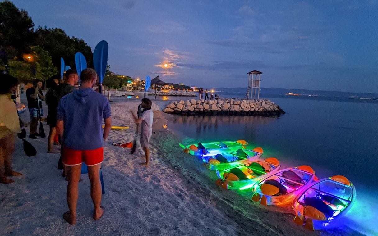 Group preparing for SUP Glow Tour in Split with illuminated kayaks on the beach at dusk.