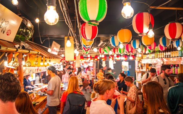 Crowded Nishiki Market in Kyoto with colorful lanterns and people enjoying street food.
