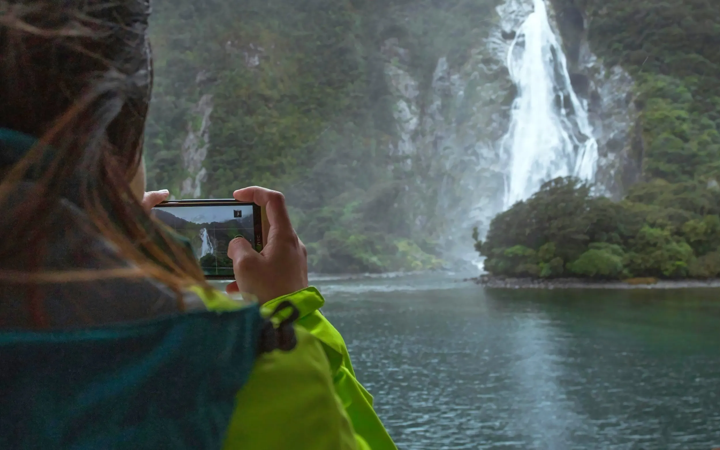 Person photographing waterfall at Milford Sound, New Zealand, during scenic cruise.