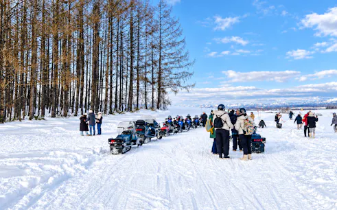 Snowmobiles and visitors at Chiyoda Snow Park, Hokkaido, with trees and mountains in the background.