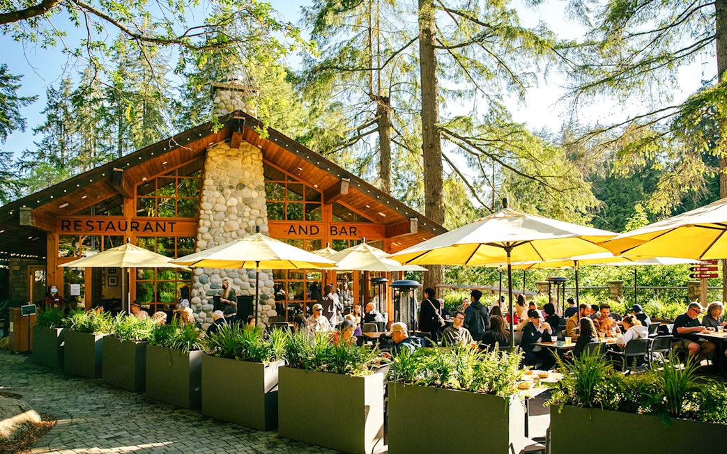 Outdoor dining area at Capilano Suspension Bridge Park with people enjoying meals under yellow umbrellas.