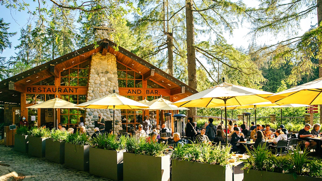 Outdoor dining area at Capilano Suspension Bridge Park with people enjoying meals under yellow umbrellas.