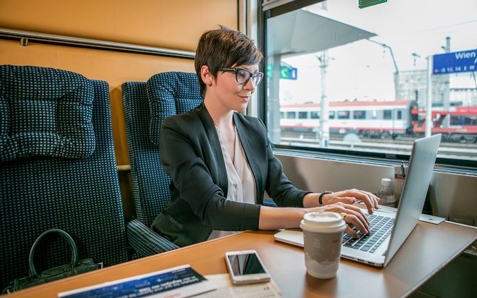 Professional woman working on laptop in first class train seat, Vienna station visible outside.