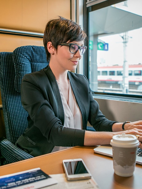 Professional woman working on laptop in first class train seat, Vienna station visible outside.