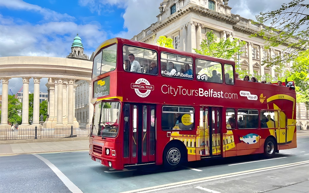 Red double-decker bus on Belfast hop-on hop-off tour passing historic building.