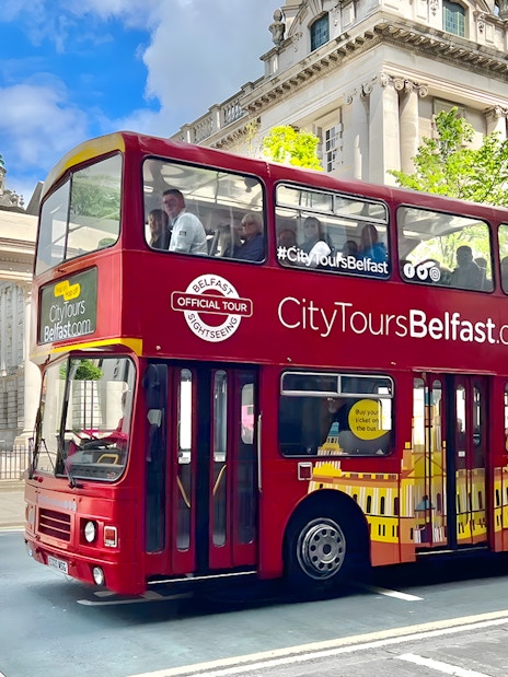 Red double-decker bus on Belfast hop-on hop-off tour passing historic building.