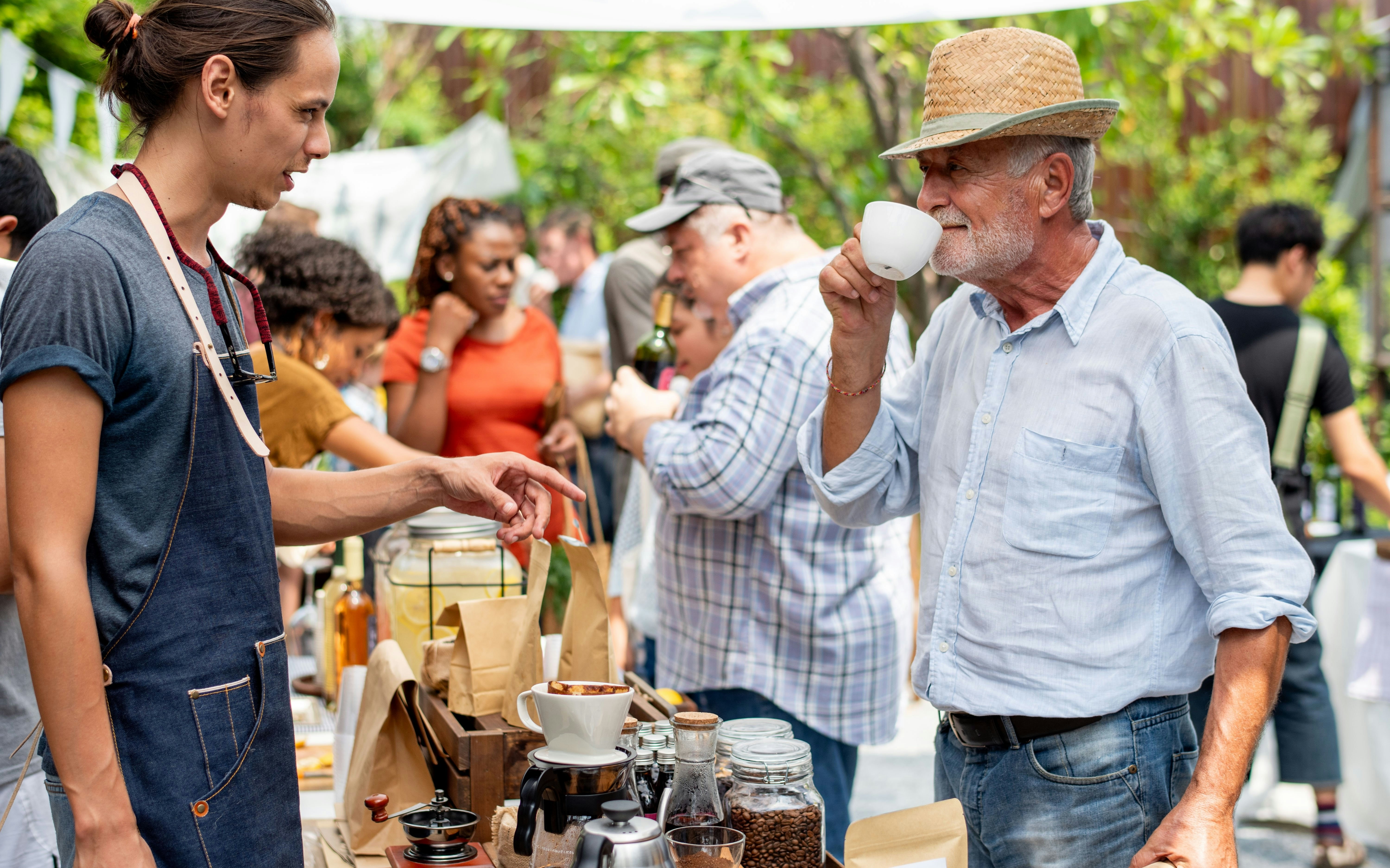 Outdoor market coffee tasting with vendor and visitor sampling brew.