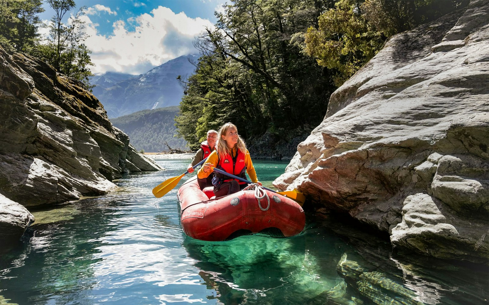 Two people paddling a red inflatable kayak on Dart River, surrounded by rocky cliffs and forest.