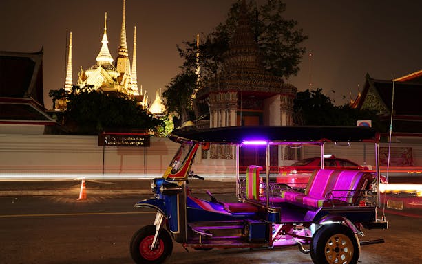 Tuk-tuk parked near illuminated temple spires in Bangkok at night.