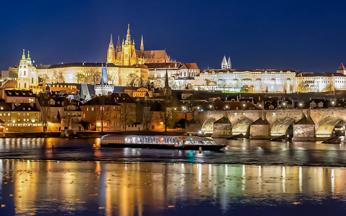 Open-top glass boat on Vltava River with Prague Castle and Charles Bridge illuminated at night.