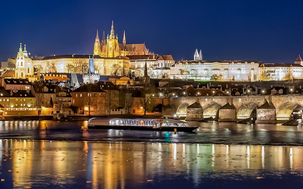 Open-top glass boat on Vltava River with Prague Castle and Charles Bridge illuminated at night.