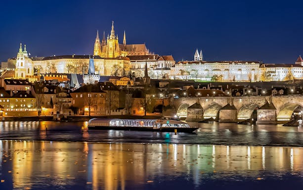 Open-top glass boat on Vltava River with Prague Castle and Charles Bridge illuminated at night.