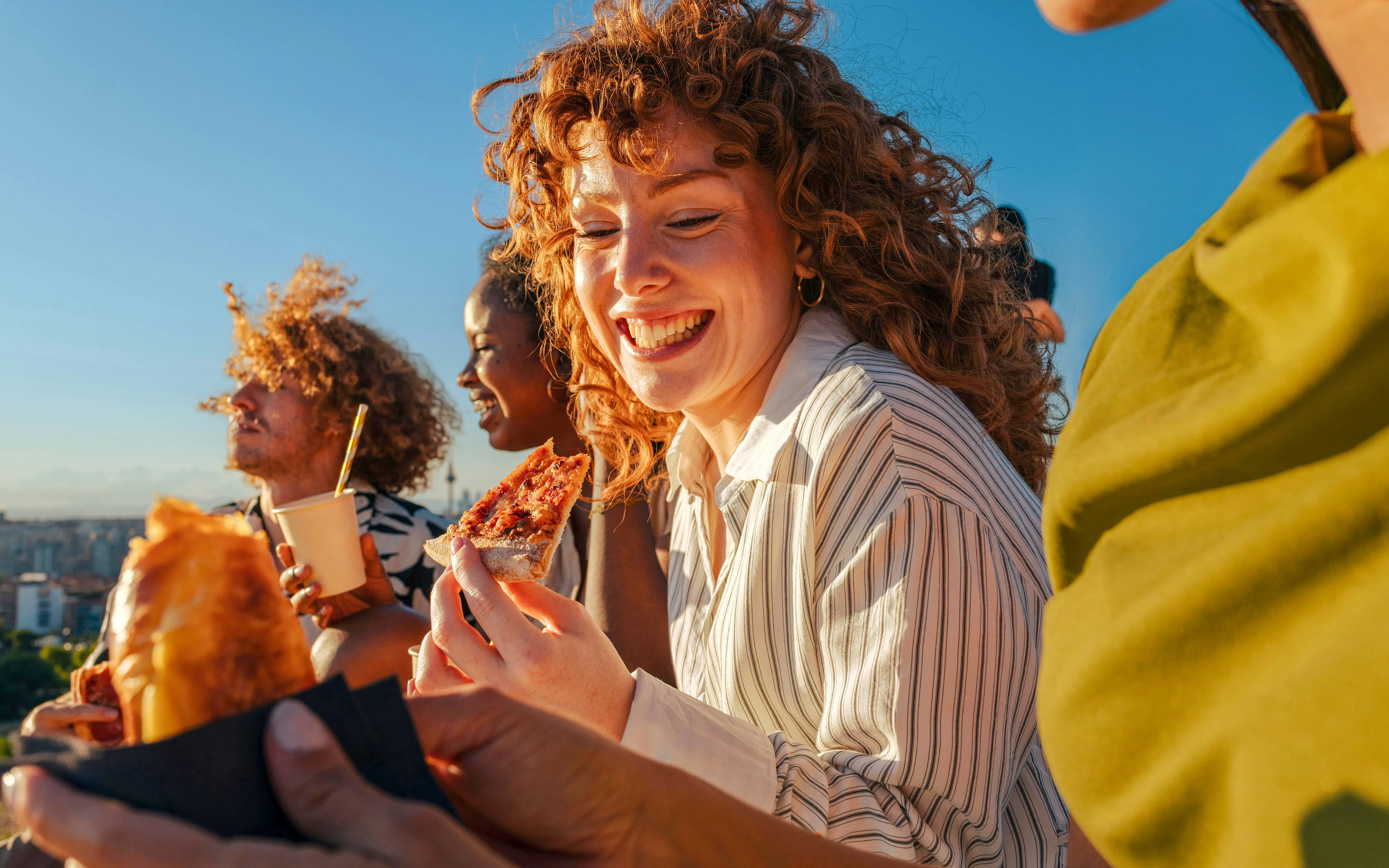 Friends enjoying pizza outdoors with city view in the background.