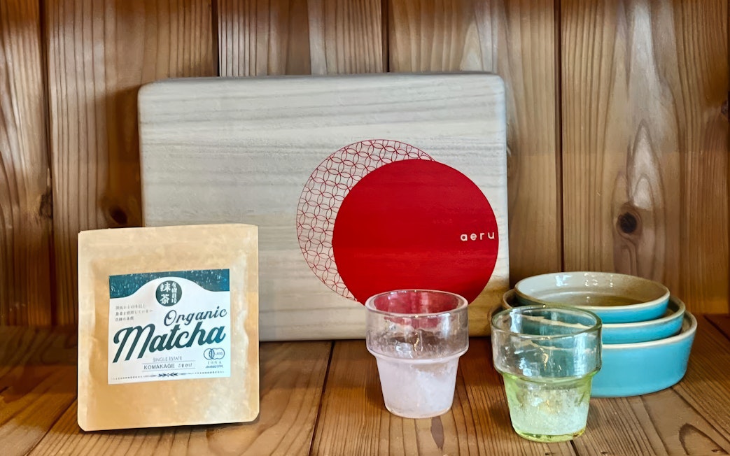 Organic matcha package with glass cups and ceramic bowls on a wooden shelf.
