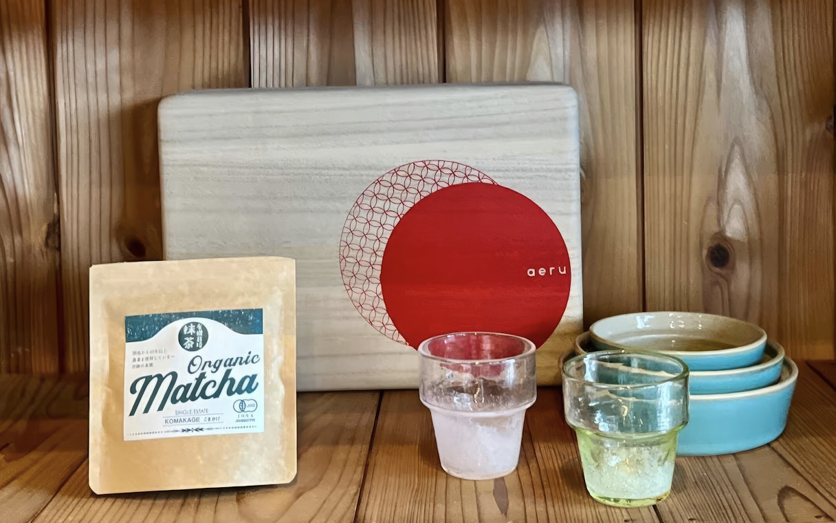 Organic matcha package with glass cups and ceramic bowls on a wooden shelf.