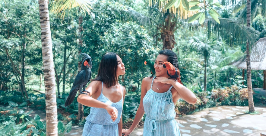 Two tourists with parrots at Lombok Wildlife Park, surrounded by lush greenery.