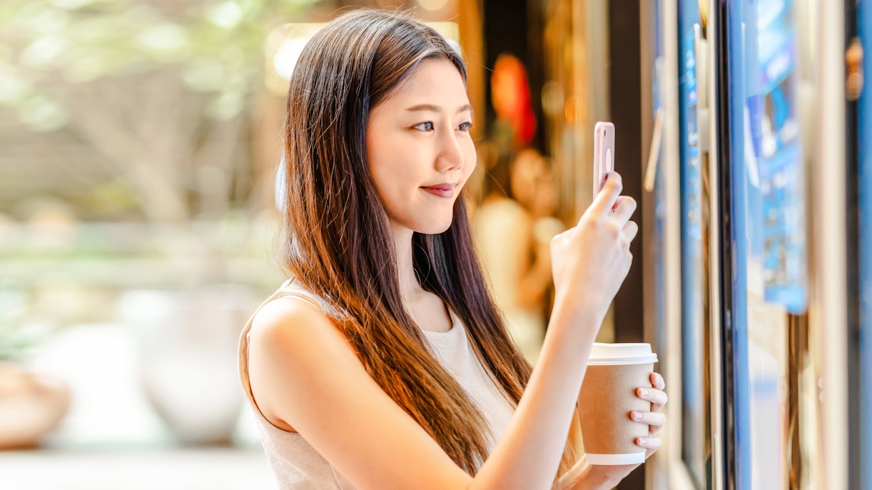 Young woman using smartphone at ticket machine in department store.