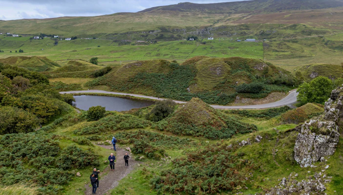 Visitors walking along a path in Fairy Glen, Isle of Skye, surrounded by lush green hills and a small pond.