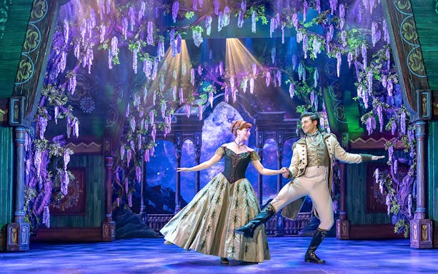 Characters dancing on stage in Disney FROZEN musical with floral backdrop.