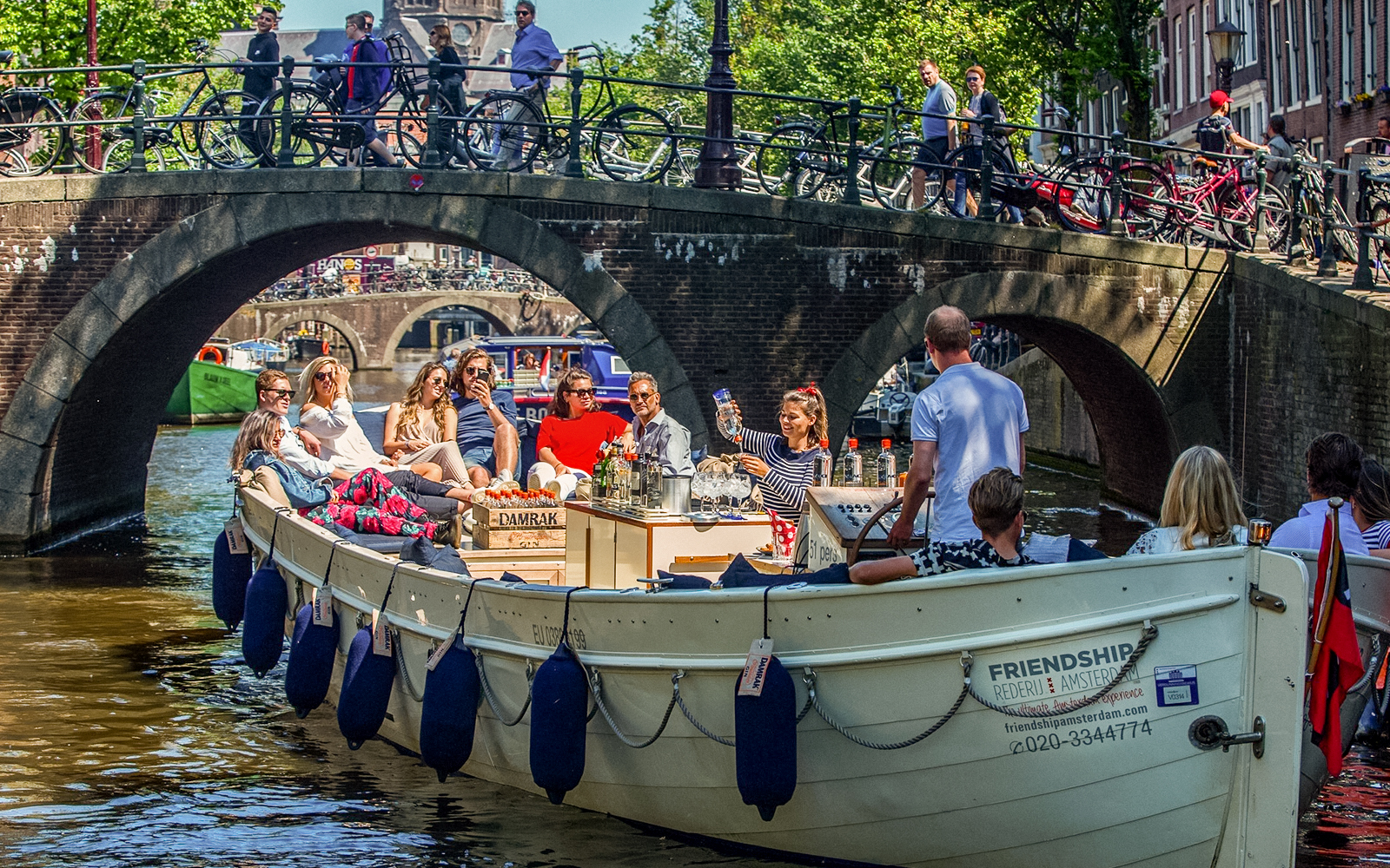 Luxury open boat cruise on Amsterdam canal under a bridge with passengers enjoying drinks.