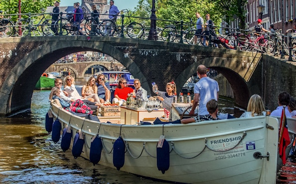 Luxury open boat cruise on Amsterdam canal under a bridge with passengers enjoying drinks.