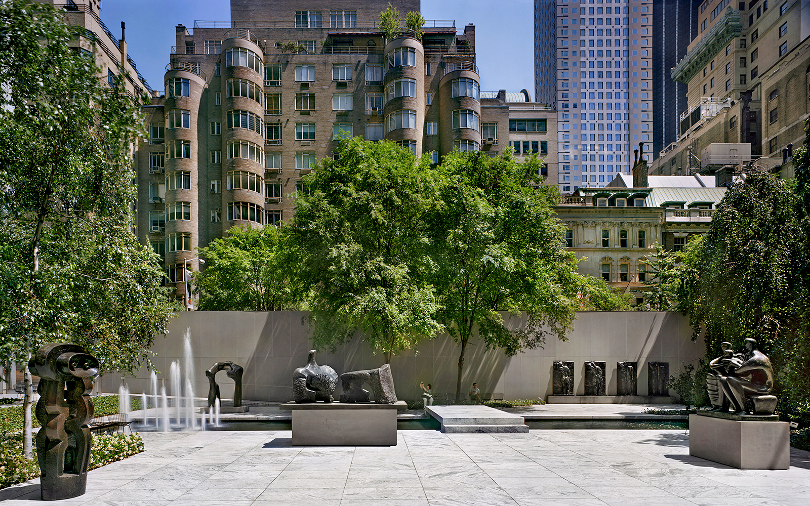 Sculpture garden with modern art pieces and fountains, surrounded by urban buildings.