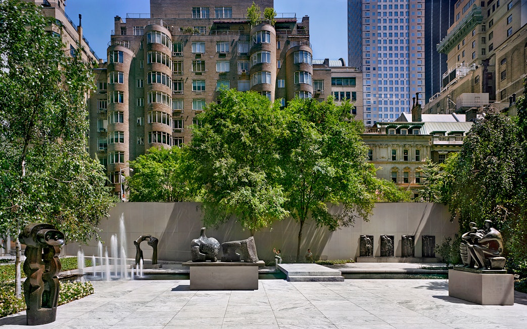 Sculpture garden with modern art pieces and fountains, surrounded by urban buildings.