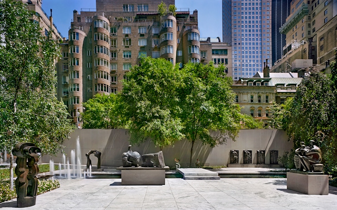 Sculpture garden with modern art pieces and fountains, surrounded by urban buildings.