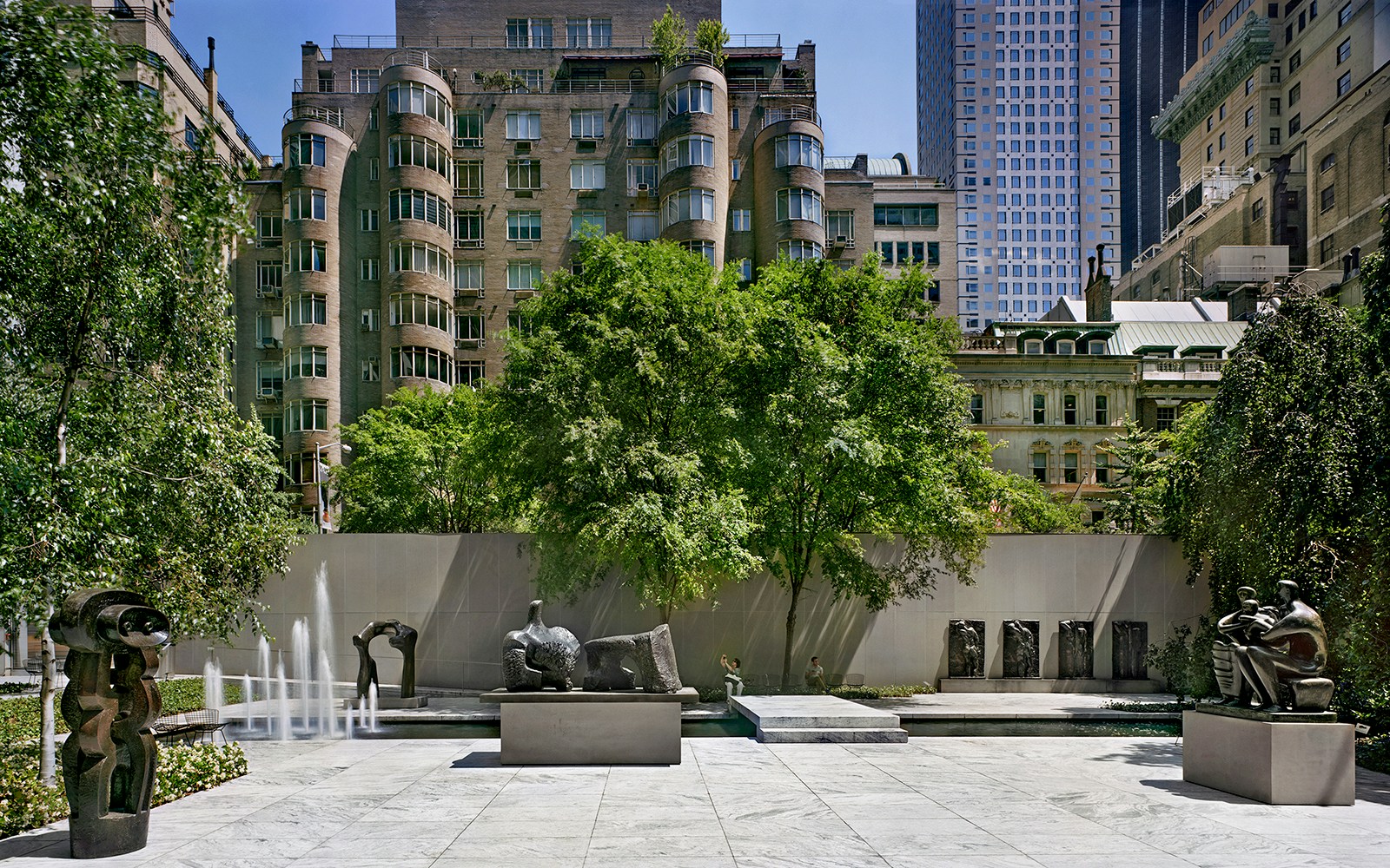 Sculpture garden with modern art pieces and fountains, surrounded by urban buildings.