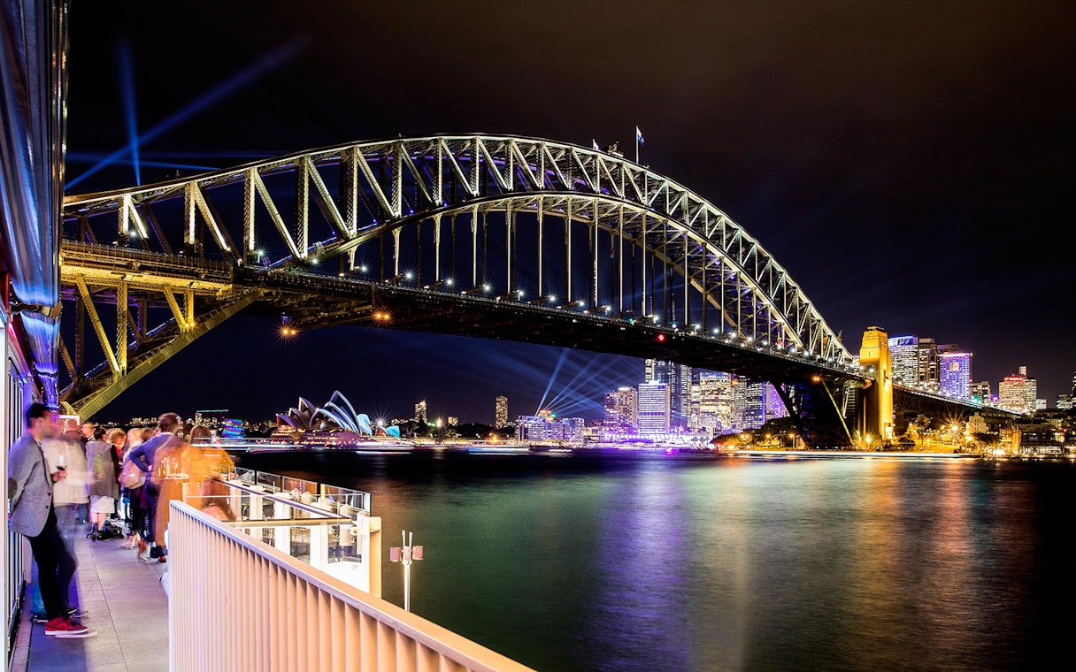 Sydney Harbour Bridge illuminated at night with city skyline and Opera House in view.